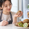 キウイを手に取って食べ頃を確認する日本人女性と、皿に盛った果実や冷蔵庫の保存容器